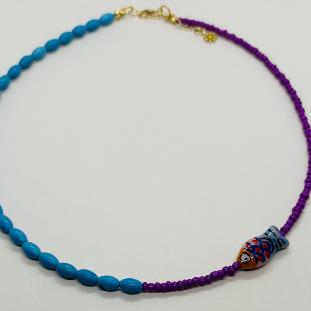 Handcrafted necklace with blue and purple beads and a fish-shaped charm on a light gray background